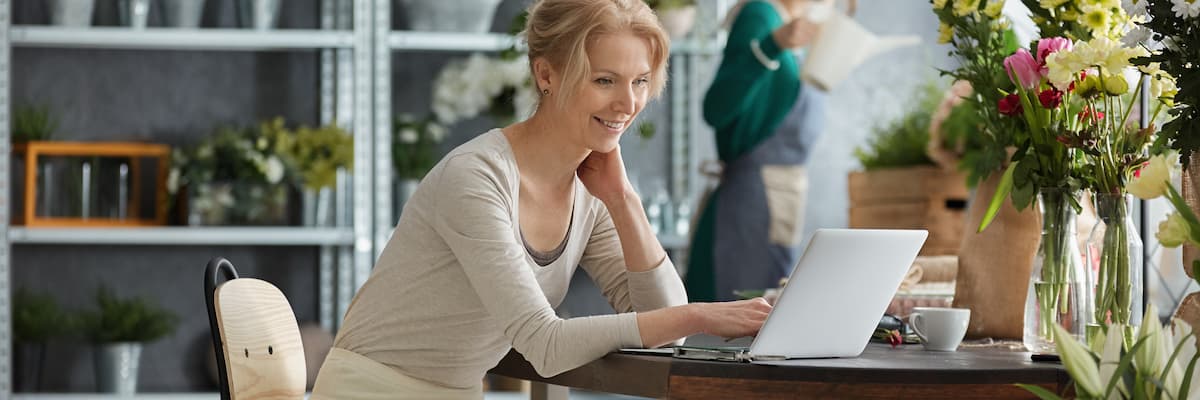 Une femme est assise à une table sur laquelle se trouve un vase de fleurs et regarde joyeusement son ordinateur portable pour comparer les logiciels de numérisation et de reconnaissance.